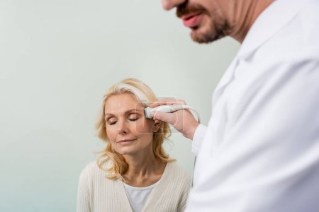 verschwommener Arzt macht Kopf-Ultraschall einer blonden Frau, die mit geschlossenen Augen sitzt