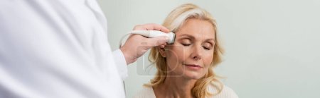 mujer rubia con los ojos cerrados cerca médico borrosa haciendo ultrasonido de su cabeza, pancarta