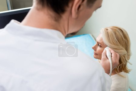 médico borroso haciendo ultrasonido de cabeza de mujer madura rubia en la clínica