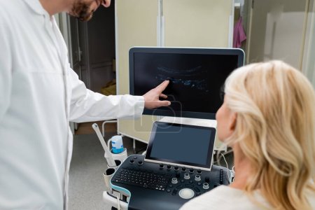 Arzt zeigt auf Bild auf Ultraschallmonitor neben blonder Frau auf verschwommenem Vordergrund