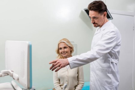 doctor pointing at monitor of ultrasound machine near smiling middle aged woman