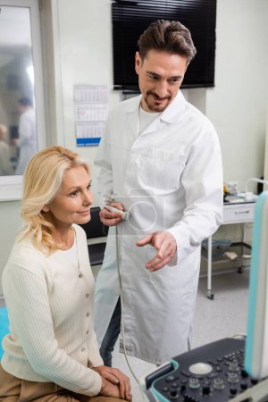 Foto de Doctor positivo apuntando al monitor de la máquina de ultrasonido cerca de la mujer rubia sonriente - Imagen libre de derechos