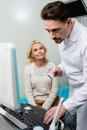 Aufmerksame Ärztin blickt auf Monitor des Ultraschallgeräts neben lächelnder Frau mittleren Alters