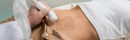 vista parcial del médico haciendo ultrasonido abdominal a la mujer en la clínica, bandera