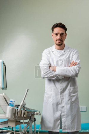 Foto de Médico serio de pie con los brazos cruzados cerca de la máquina de ultrasonido y mirando a la cámara - Imagen libre de derechos