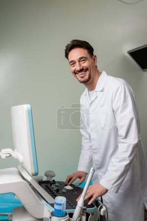 Photo for Happy doctor in white coat looking at camera while operating ultrasound machine in clinic - Royalty Free Image