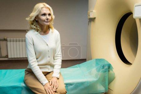 Lächelnde Frau mittleren Alters sitzt neben Computertomographen in Klinik