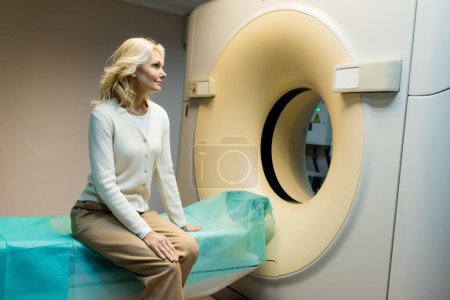 Lächelnde blonde Frau sitzt neben Computertomographen in Klinik