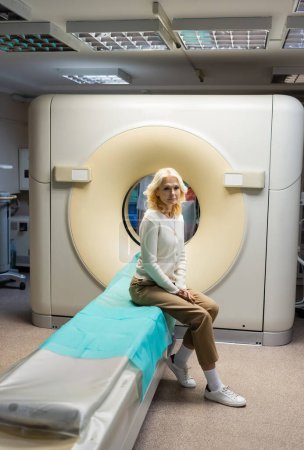 full length of blonde mature woman sitting near computed tomography machine in clinic