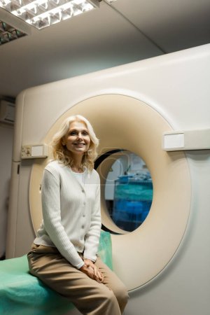 cheerful mature woman sitting near computed tomography scanner and looking at camera