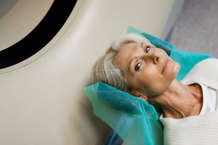 Hochwinkelaufnahme einer blonden Frau mittleren Alters, die während der Untersuchung auf dem CT-Scanner in die Kamera schaut
