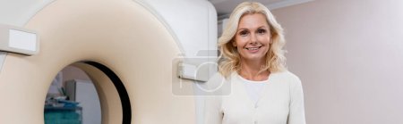 Photo for Happy blonde woman smiling at camera near ct scanner in hospital, banner - Royalty Free Image