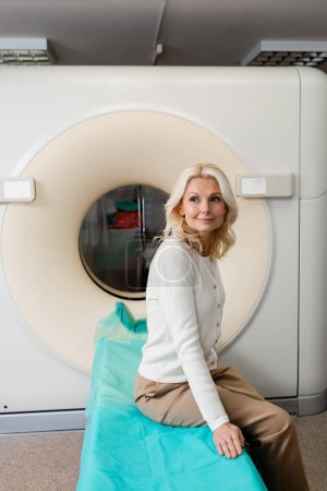 mujer madura positiva sentada cerca del escáner ct en la clínica y mirando hacia otro lado