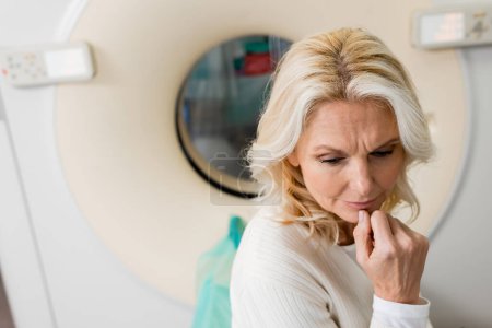 nervous middle aged woman holding hand near face near ct scanner on blurred background