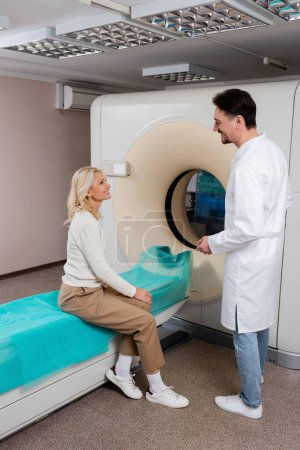 Photo for Radiologist in white coat talking to smiling mature woman sitting near computed tomography machine - Royalty Free Image