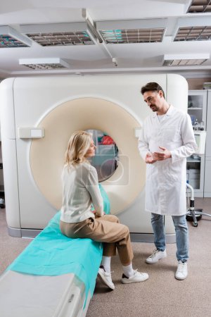 Téléchargez les photos : Radiologue pleine longueur en manteau blanc parlant à une femme blonde assise près de ct scanner à la clinique - en image libre de droit