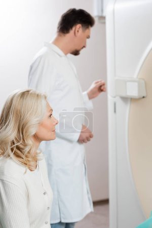 mujer rubia de mediana edad cerca del radiólogo y escáner de tomografía computarizada en primer plano borroso