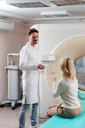 Téléchargez les photos : Médecin positif en manteau blanc parlant à une femme mature assise près d'un scanner de tomodensitométrie - en image libre de droit
