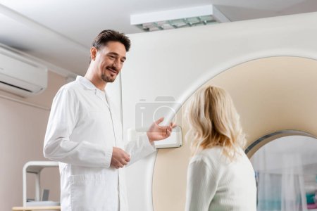 smiling doctor pointing at computed tomography scanner near blonde middle aged woman