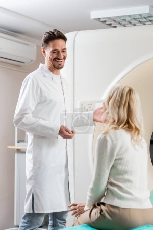 cheerful doctor in white coat talking to blonde woman near computed tomography machine