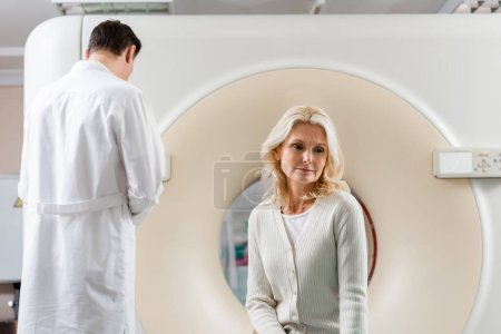 Nachdenkliche Blondine sitzt neben Scanner und Arzt im Krankenhaus