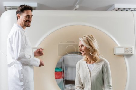 Fröhlicher Radiologe schaut lächelnde reife Frau in der Nähe von Computertomographen an