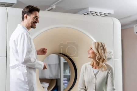 Photo for Blonde middle aged woman and positive radiologist looking at each other near ct scanner in clinic - Royalty Free Image