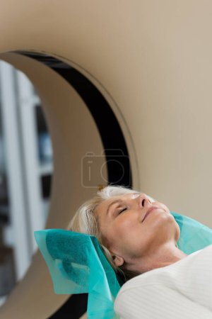 Reife Frau mit geschlossenen Augen lächelt während der Diagnostik am Computertomographen