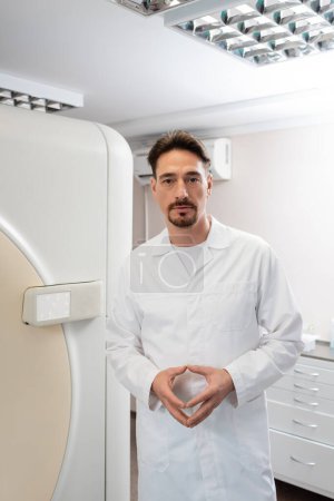 Photo for Brunette doctor in white coat looking at camera near computed tomography machine in clinic - Royalty Free Image