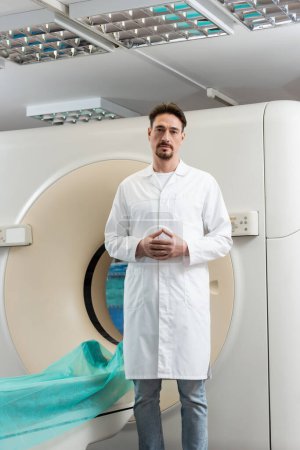 Schwerbärtiger Radiologe blickt in Klinik auf Kamera in der Nähe von Computertomographie-Scanner