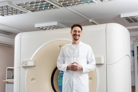 Glücklicher Radiologe im weißen Kittel steht neben Computertomographen und blickt in die Kamera