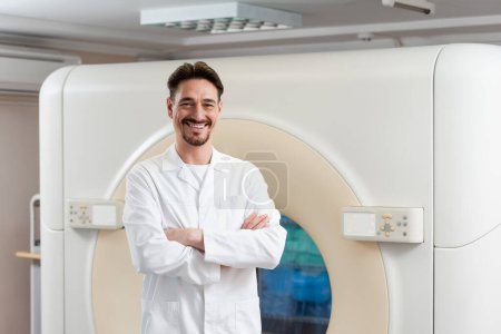 bearded physician in white coat smiling at camera near ct scanner in hospital