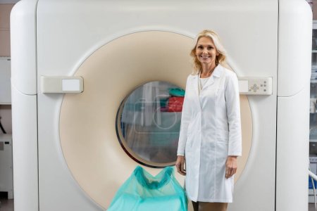 Hübsche blonde Radiologin im weißen Kittel lächelt in die Kamera neben dem Computertomographen