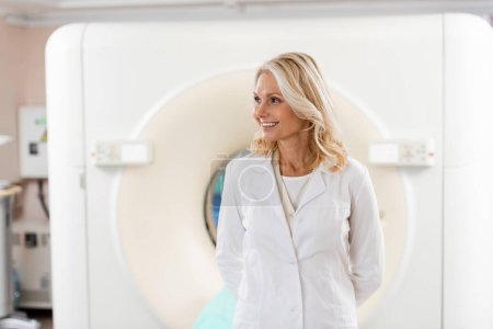 Lächelnder blonder Arzt in weißem Mantel schaut in der Nähe des Scanners im Krankenhaus weg