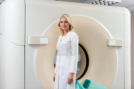 Photo for Middle aged radiologist in white coat standing near computed tomography scanner and looking at camera - Royalty Free Image