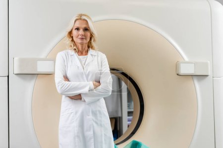 Blonder Radiologe im weißen Mantel steht mit verschränkten Armen und blickt in die Kamera neben dem Scanner
