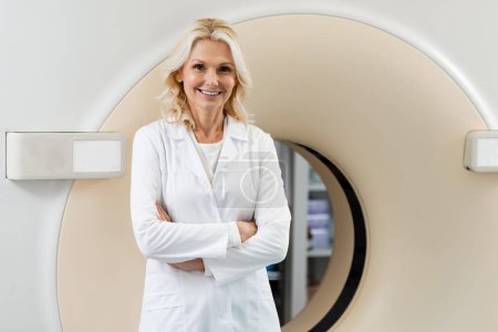Photo for Joyful physician in white coat standing with crossed arms near computed tomography scanner - Royalty Free Image