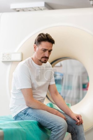 Trauriger Mann im weißen T-Shirt sitzt neben Computertomographen im Krankenhaus