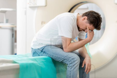 Photo for Stressed man sitting near ct scanner with bowed head and covering face with hand - Royalty Free Image