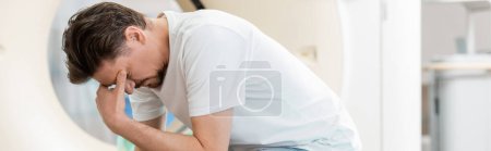 hombre frustrado cubriendo la cara mientras está sentado con la cabeza inclinada cerca del escáner de tomografía computarizada, pancarta