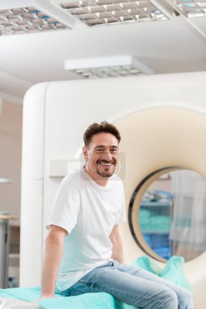 cheerful brunette man in white t-shirt sitting near ct scanner and smiling at camera