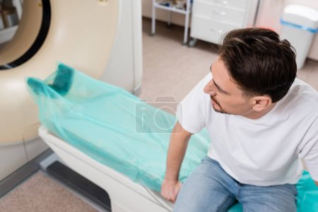 high angle view of brunette man in white t-shirt sitting near ct scanner in clinic