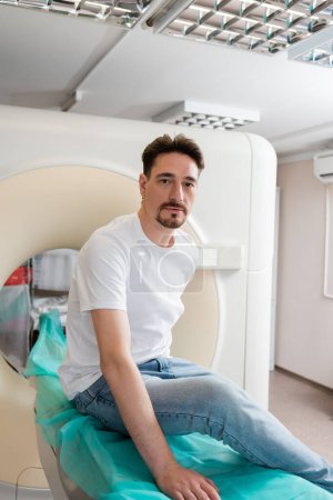 adult man in t-shirt and jeans sitting near computed tomography scanner and looking at camera