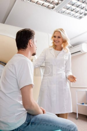 Téléchargez les photos : Radiologue d'âge moyen souriant parlant à un homme brune près d'un scanner de tomodensitométrie - en image libre de droit