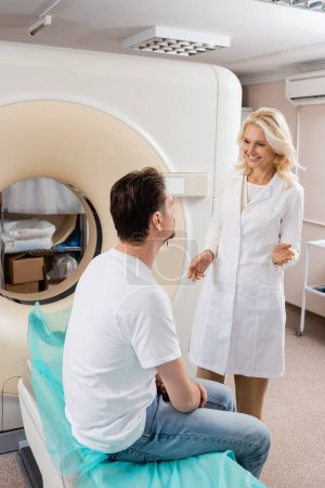 Téléchargez les photos : Radiologue blonde souriante parlant à un homme brune assis près d'un scanner de tomodensitométrie - en image libre de droit