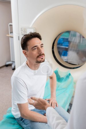 Photo for Man sitting near computed tomography machine and looking at doctor on blurred foreground - Royalty Free Image