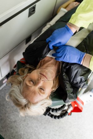 Vista superior de paramédico quitándose la chaqueta de la mujer de mediana edad inconsciente en el vehículo de emergencia 
