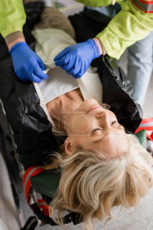 Photo for Paramedic taking off clothes from unconscious mature woman in emergency vehicle - Royalty Free Image