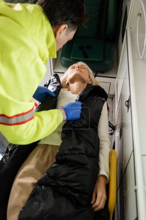 Photo for Blurred paramedic taking off jacket from unconscious middle aged woman in emergency vehicle - Royalty Free Image