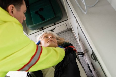 Vista de ángulo alto de paramédico borroso dando primeros auxilios a la mujer madura inconsciente en el vehículo de emergencia 